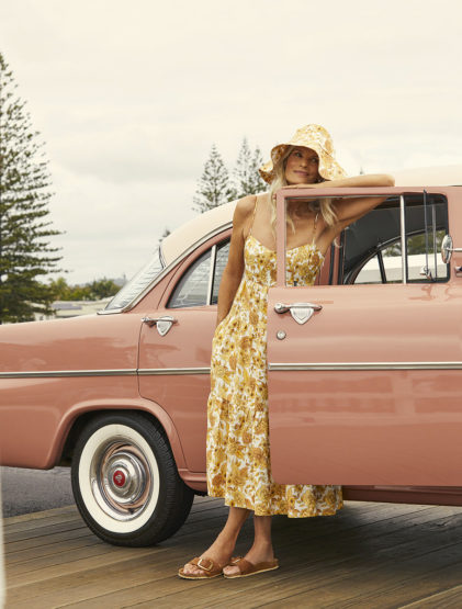 Woman in floral dress leans against a pink vintage car with trees in the background. A light-hearted outdoor fashion scene.
