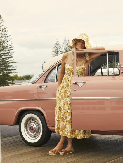 Woman in floral dress leans against a pink vintage car with trees in the background. A light-hearted outdoor fashion scene.