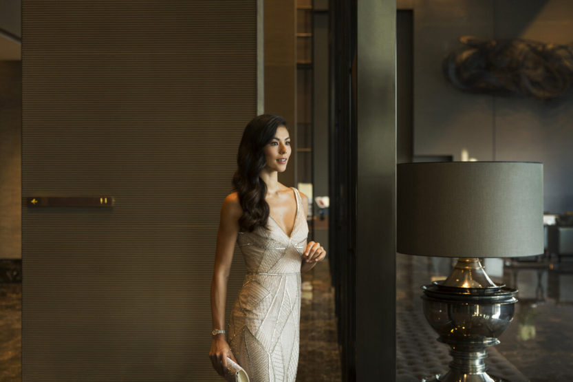 Elegant woman in a beaded gown stands in a luxurious interior, featuring warm lighting, a neutral color palette, and sophisticated decor elements.