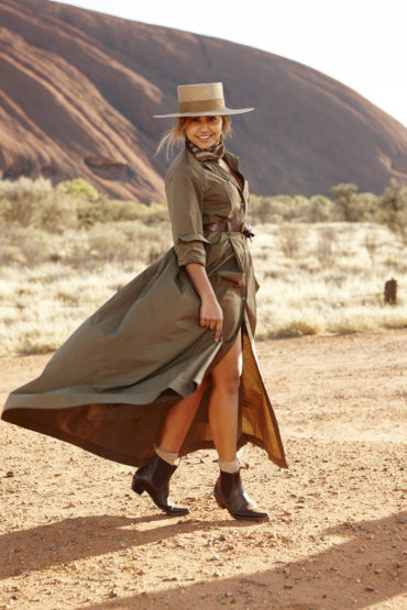 Adult female posing in an olive-green coat dress and cowboy boots, standing in front of a large red rock formation in the desert.
