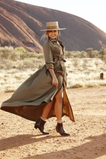 Adult female posing in an olive-green coat dress and cowboy boots, standing in front of a large red rock formation in the desert.