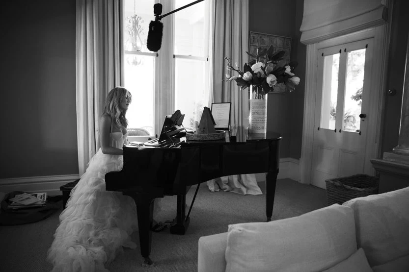 Woman in strapless gown playing grand piano in a room with natural light from large windows and neutral decor in black and white.