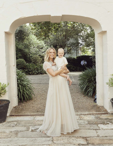 A woman in a long dress holds a baby in an outdoor setting, framed by an arched stone entrance, surrounded by lush greenery.