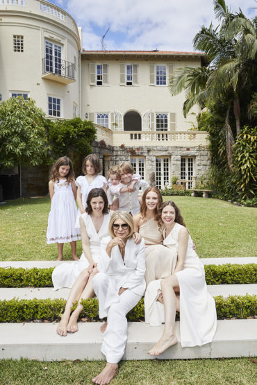 Family portrait in front of a large house. Seven females and one male in white clothing. Green lawn and trees surround the scene.