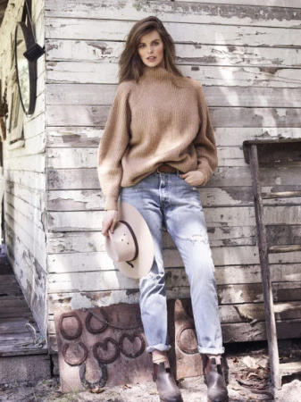 Woman in beige turtleneck sweater and light blue jeans leaning against a weathered wooden rustic wall