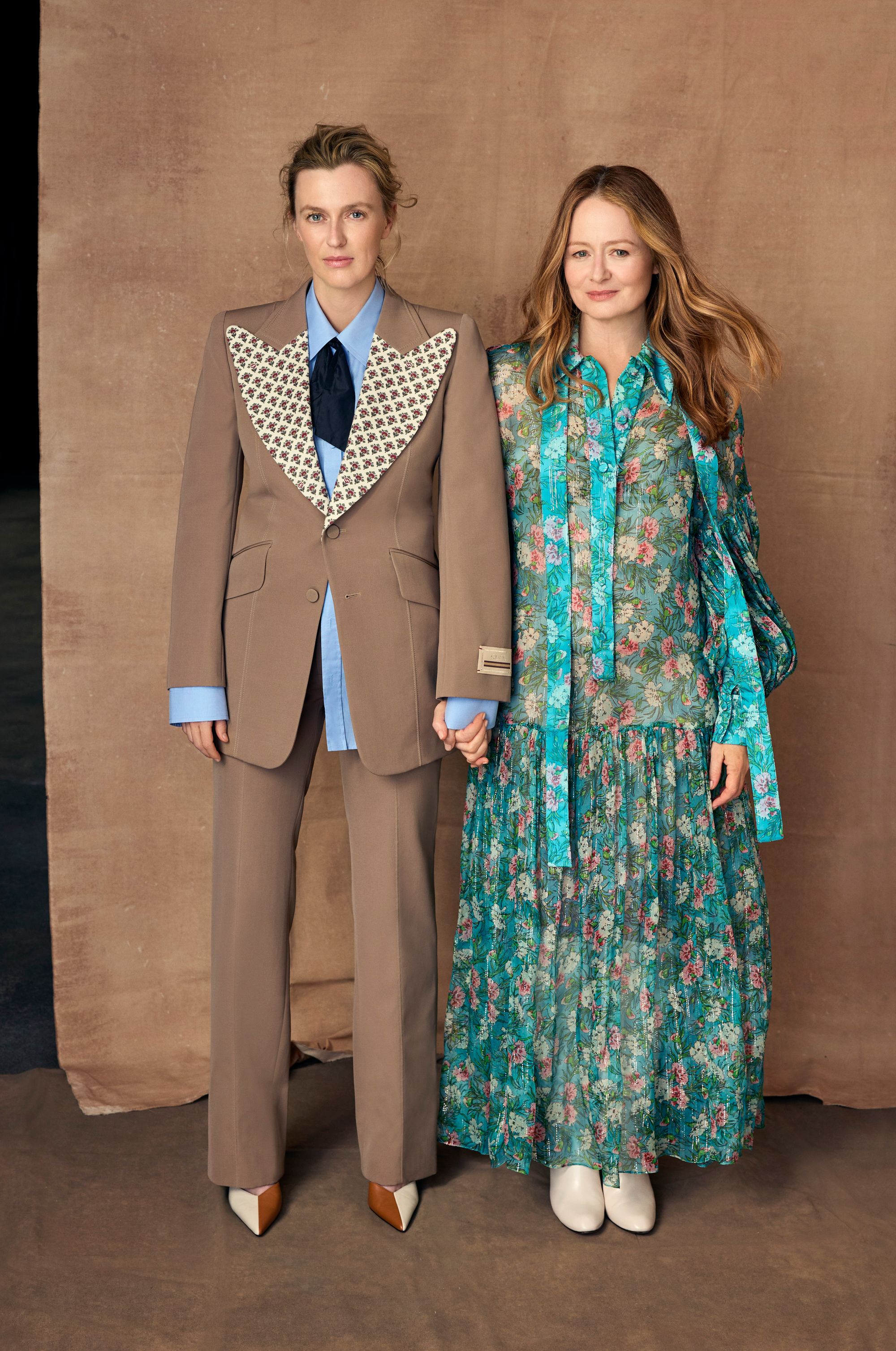 Two women pose in stylish outfits against a neutral backdrop. One in a brown suit with patterned lapels, the other in a floral dress.