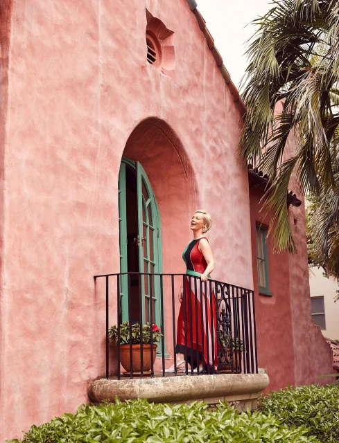 Woman in formal dress standing on a wrought-iron balcony with a peach colored stucco building and palm trees in the background.