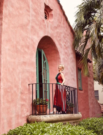 Woman in formal dress standing on a wrought-iron balcony with a peach colored stucco building and palm trees in the background.