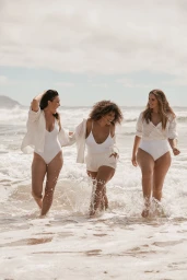 Three women in white swimwear wading through ocean surf at the beach