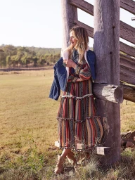 Woman in a striped maxi dress and denim jacket leaning against a wooden post in an open field styled by Mattie Cronan