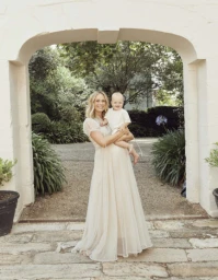 Woman in a long cream sheer dress holding a toddler beneath a stone archway in a garden