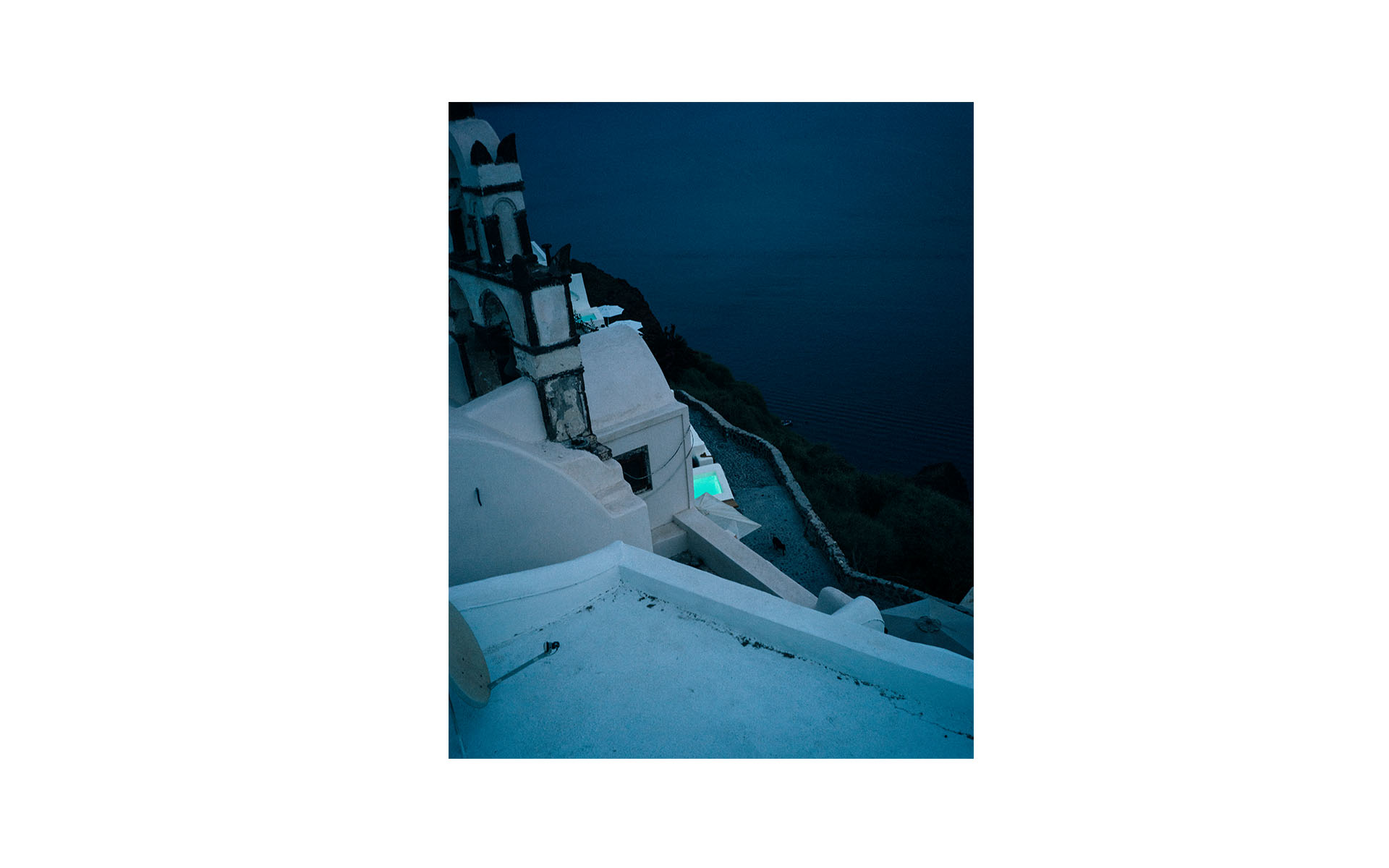 High-angle view of white buildings on a cliffside, with a glimpse of the dark blue sea in the background under a dim, night sky.