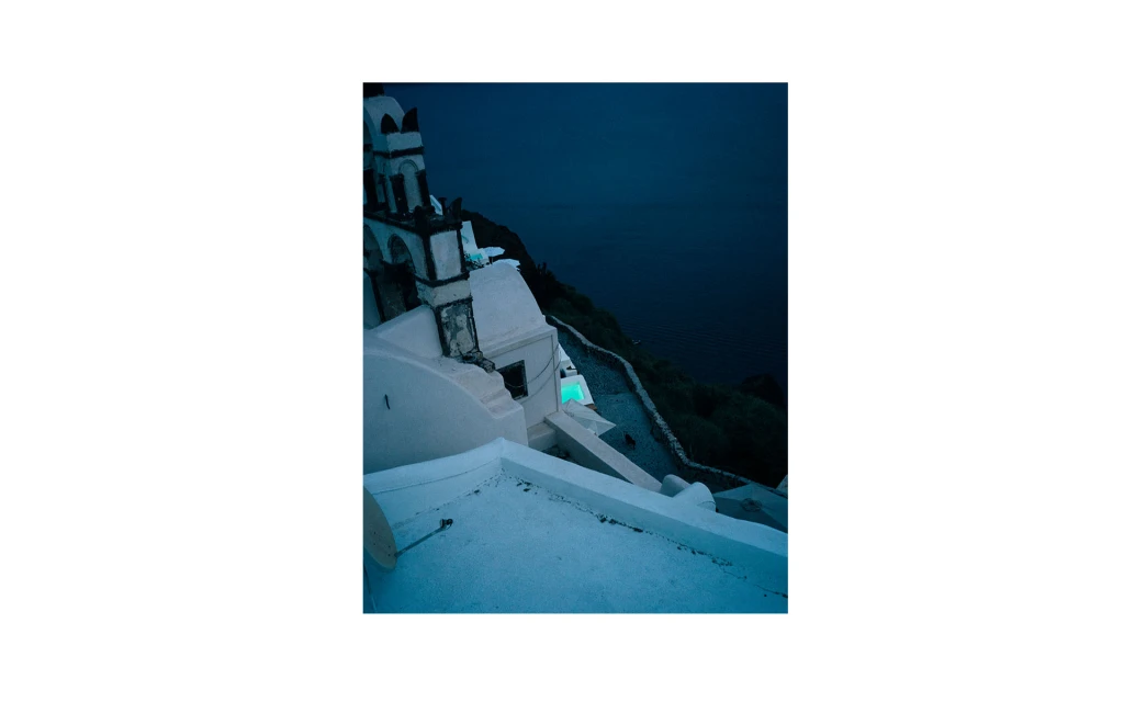 High-angle view of white buildings on a cliffside, with a glimpse of the dark blue sea in the background under a dim, night sky.