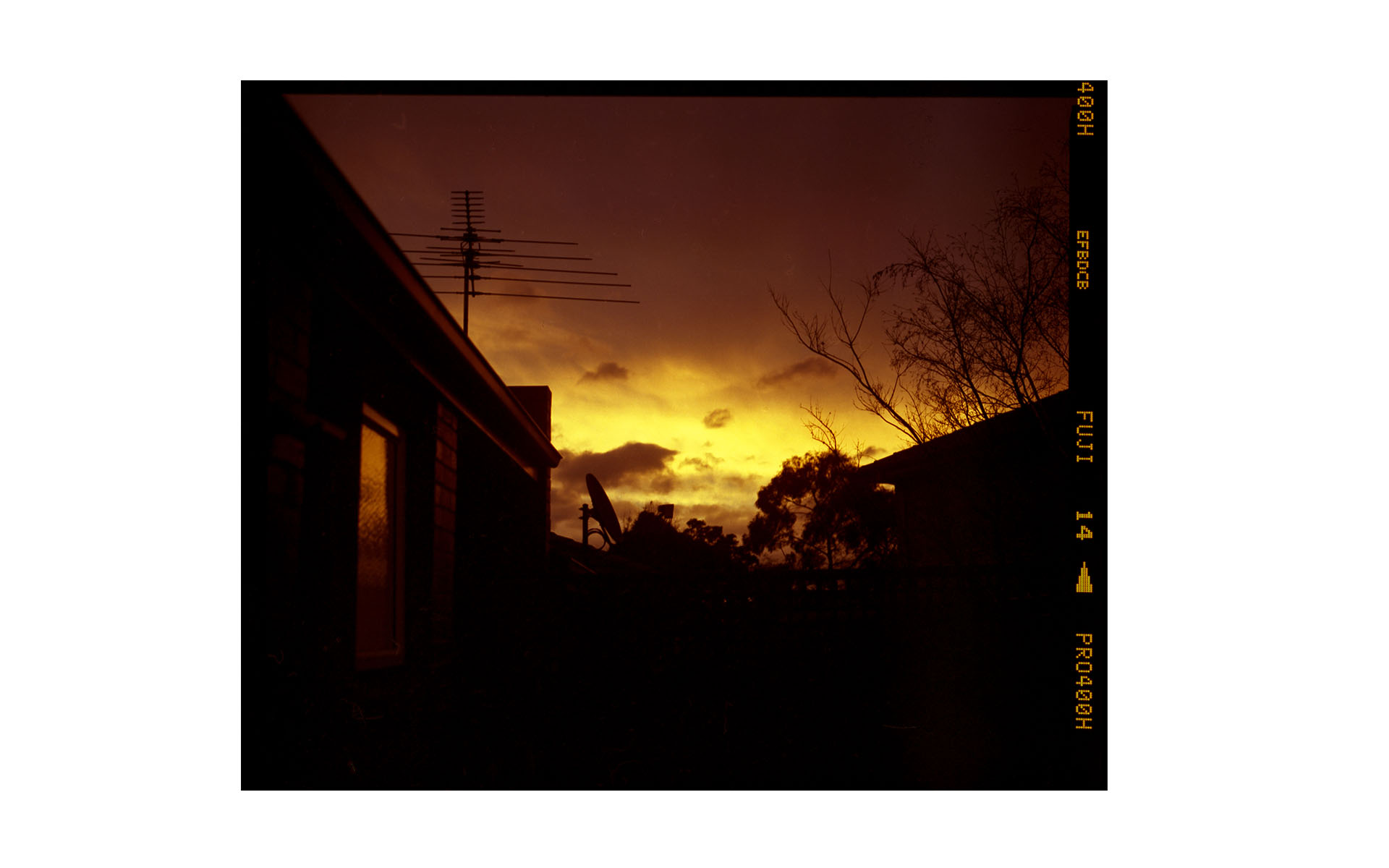 Sky at sunset with silhouettes of houses and an antenna. The sky has colors from gold to orange and the photo has a film border.