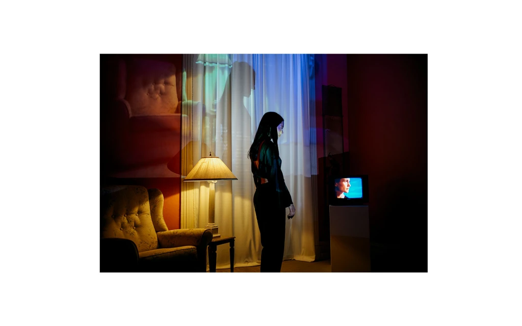 An adult woman in dark clothing stands in a room with an old television, lit by colorful lights and accented by vintage furniture.