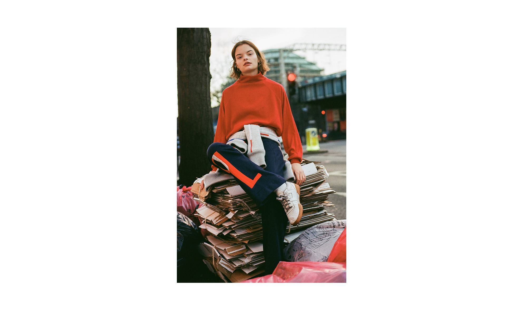 A woman with a red sweater and athletic pants sits on a stack of cardboard boxes on an urban street with a tree and overpass in the background.