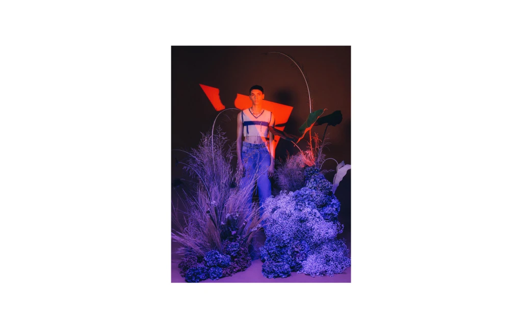 Full shot of an adult in a crop top and jeans standing amongst floral plants, grasses, and geometric structures in a studio.