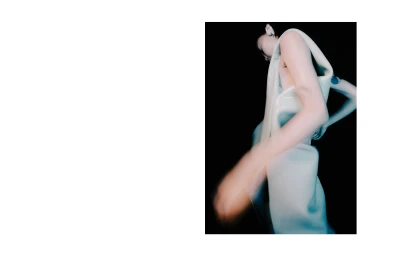A model in a sculptural white sleeveless garment with a blurred arm motion against a stark black background.