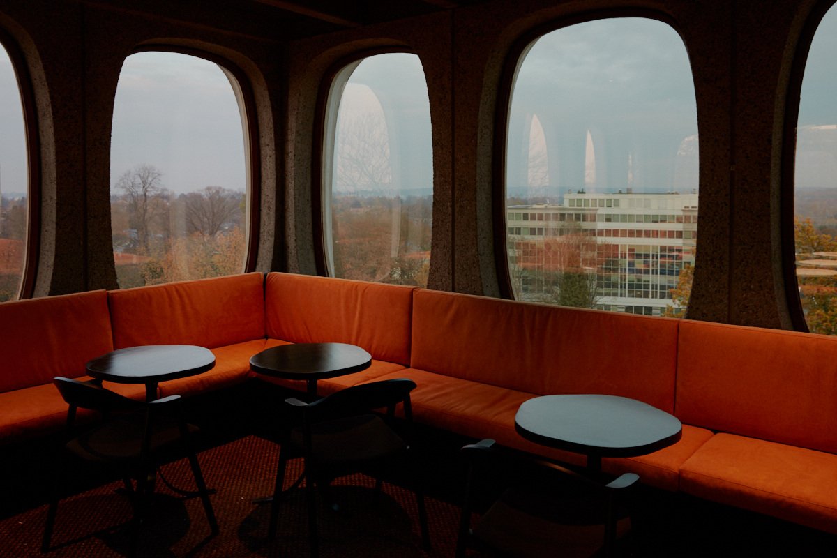 Interior view with orange cushioned seating and round tables, set against a backdrop of large, arched windows showing a cityscape.