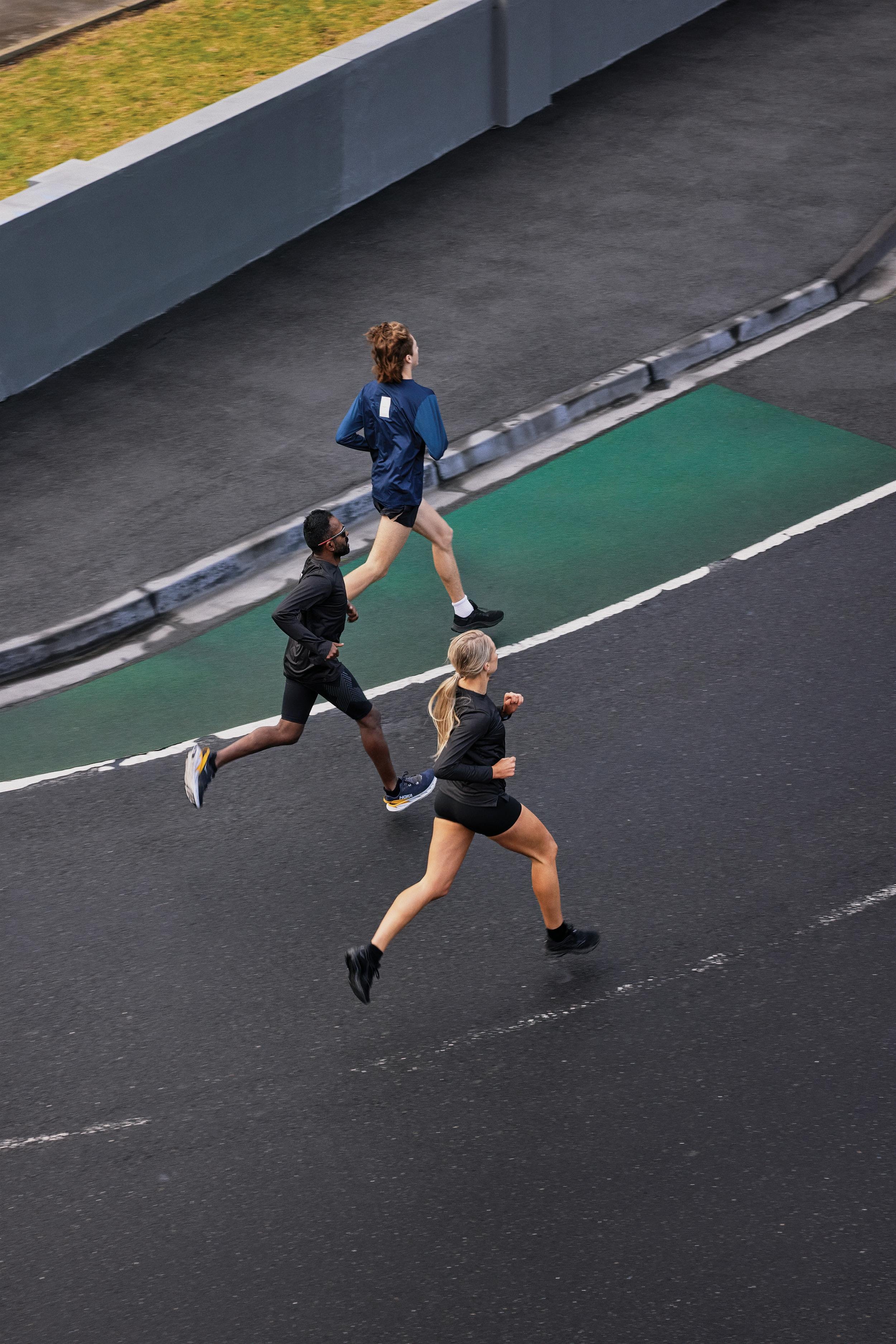 Three athletes run on an asphalt surface near a sidewalk and grass area. They are wearing athletic clothing and shoes. High-angle view.