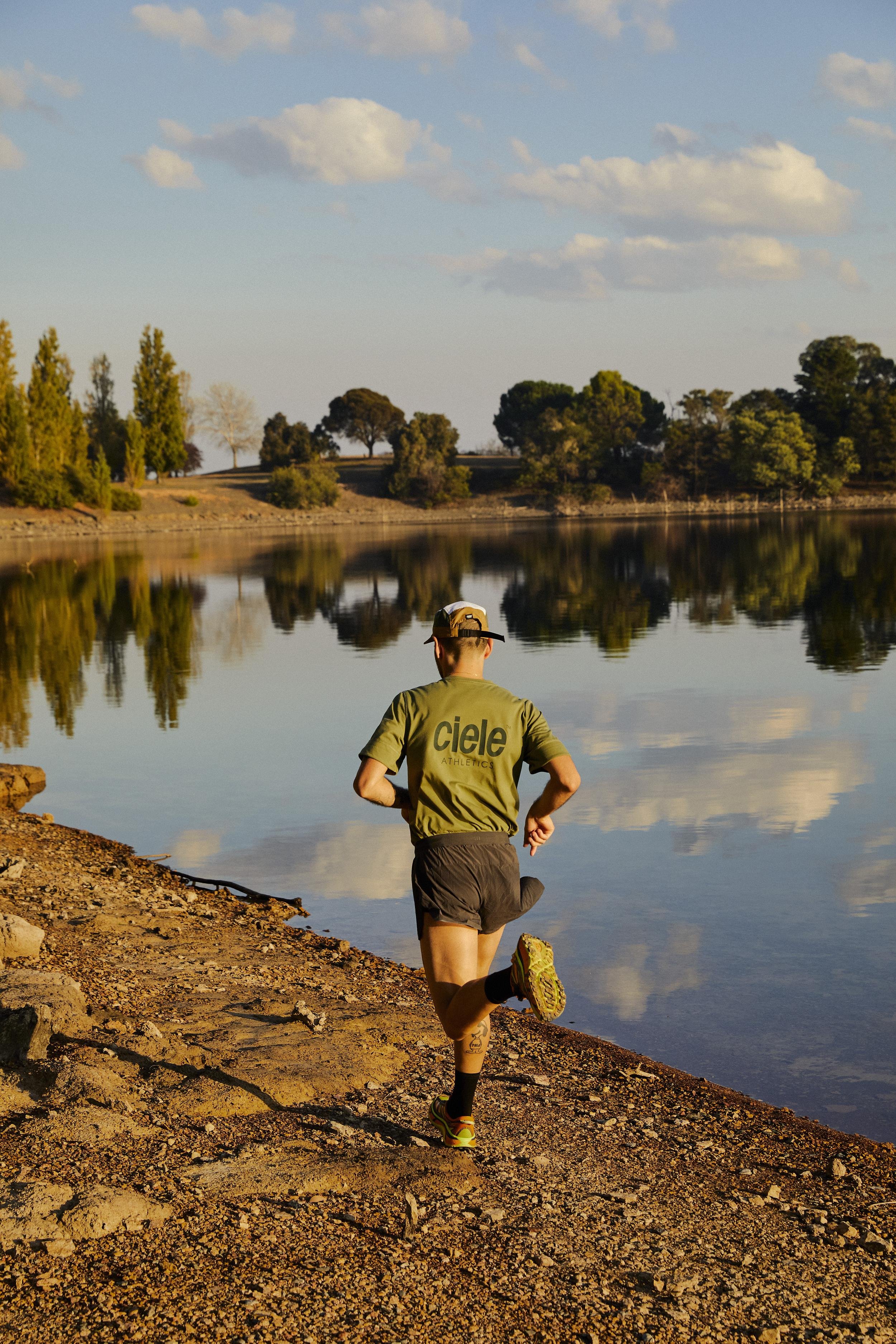 Athletic man in activewear runs along rocky lakeside trail at sunset with reflections in water, trees, and partly cloudy blue sky.
