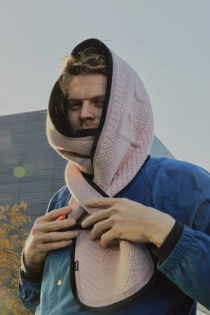 Adult male wearing pink hooded scarf outdoors against the sky and a modern architectural building in medium portrait shot.