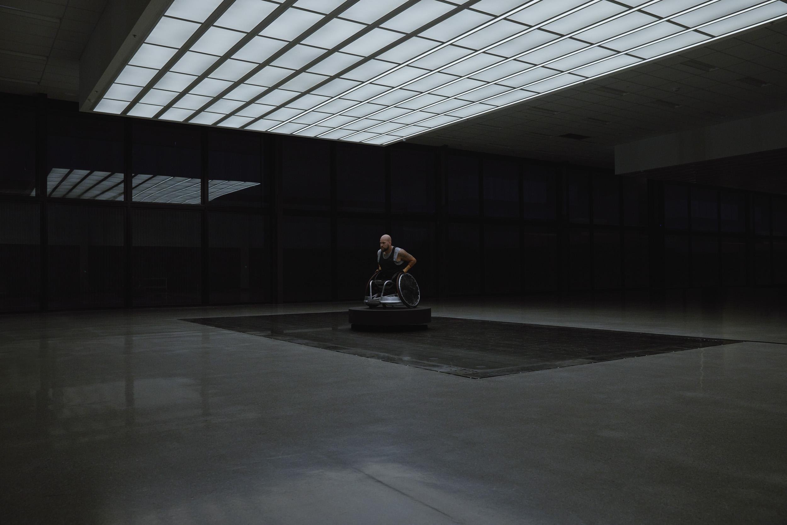 An adult male with a shaved head, seated in a wheelchair on a circular platform, in a vast, dimly lit interior space.