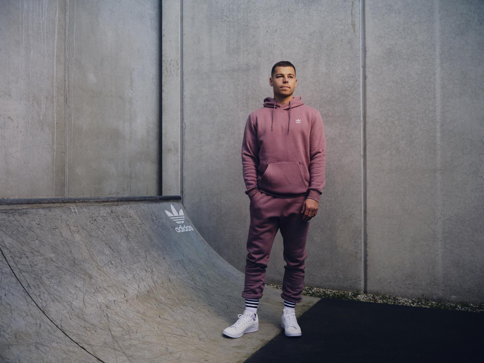 Man in a mauve tracksuit and white sneakers stands outdoors against a concrete wall in natural light, showcasing casual style and Adidas branding.