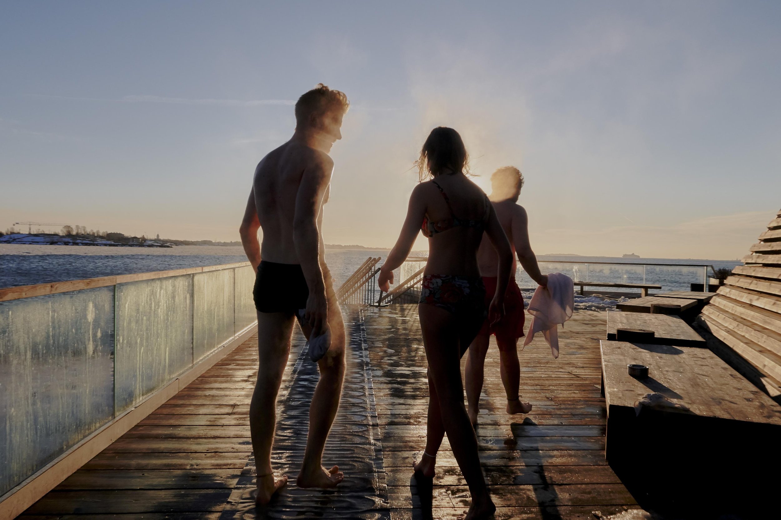 Three people in swimwear stand on a wooden dock near a body of water with a distant shoreline and clear sky at daytime.
