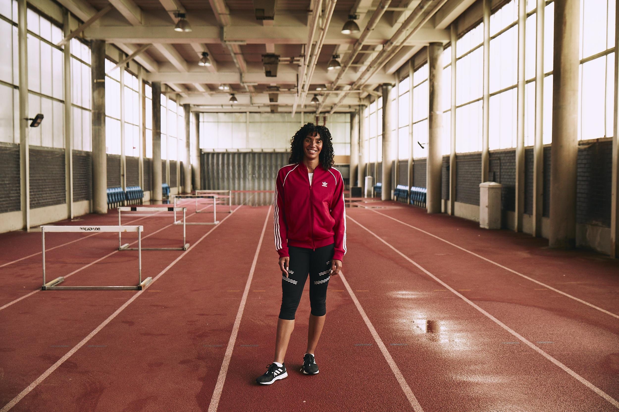 Female athlete stands confidently on an indoor running track wearing a red Adidas tracksuit and black leggings, with hurdles in the background.