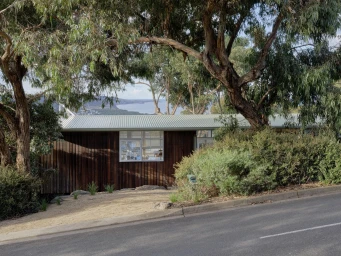 Single-story timber-clad house nestled among eucalyptus trees with a distant view of the ocean, photographed from a roadside.