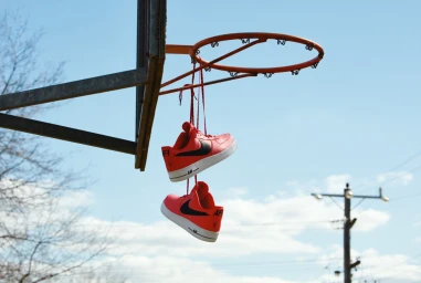 Red sneakers hanging by their laces from an outdoor orange basketball hoop against a clear blue sky background.