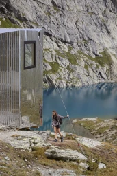 Hiker holding a guideline near a mirrored metal cabin with a mountain lake background