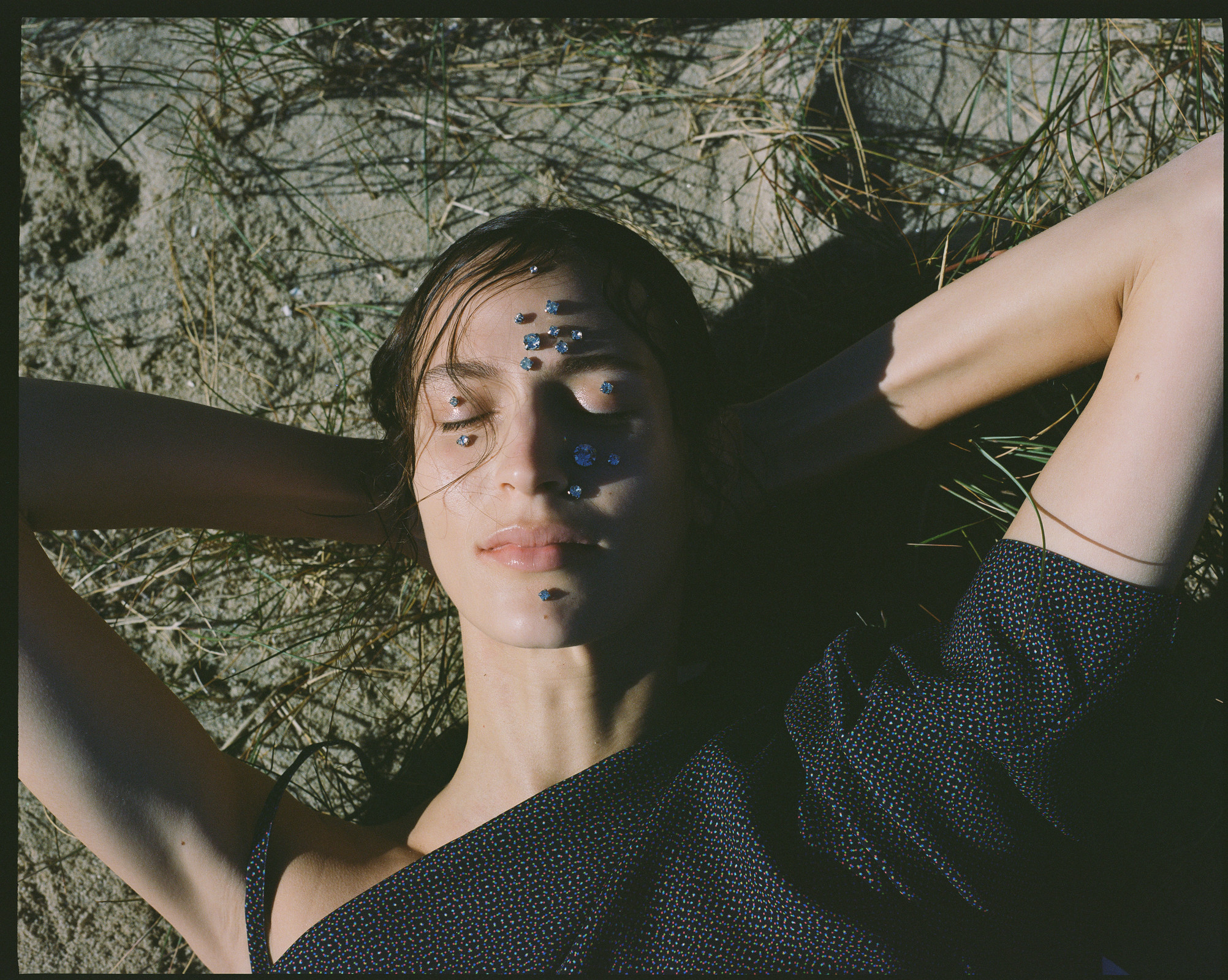 Woman with jewels on face lies in grass in dark grey top. Natural light, medium shot, outdoor environment, conceptual fashion.