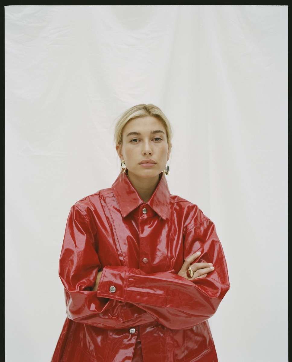 Blonde woman with fair skin poses in a shiny red jacket against a white backdrop, wearing gold hoop earrings and a ring.