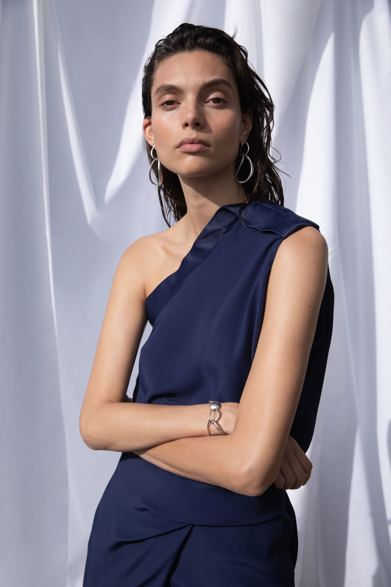 Portrait of an adult woman in a one-shoulder navy blue dress with jewelry, standing indoors against a white draped background.