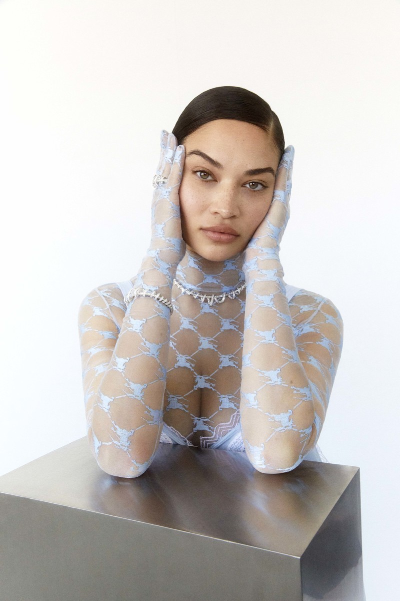 A fashion portrait features an adult woman with hands on her face, wearing light blue patterned gloves and jewelry in a bright indoor setting.