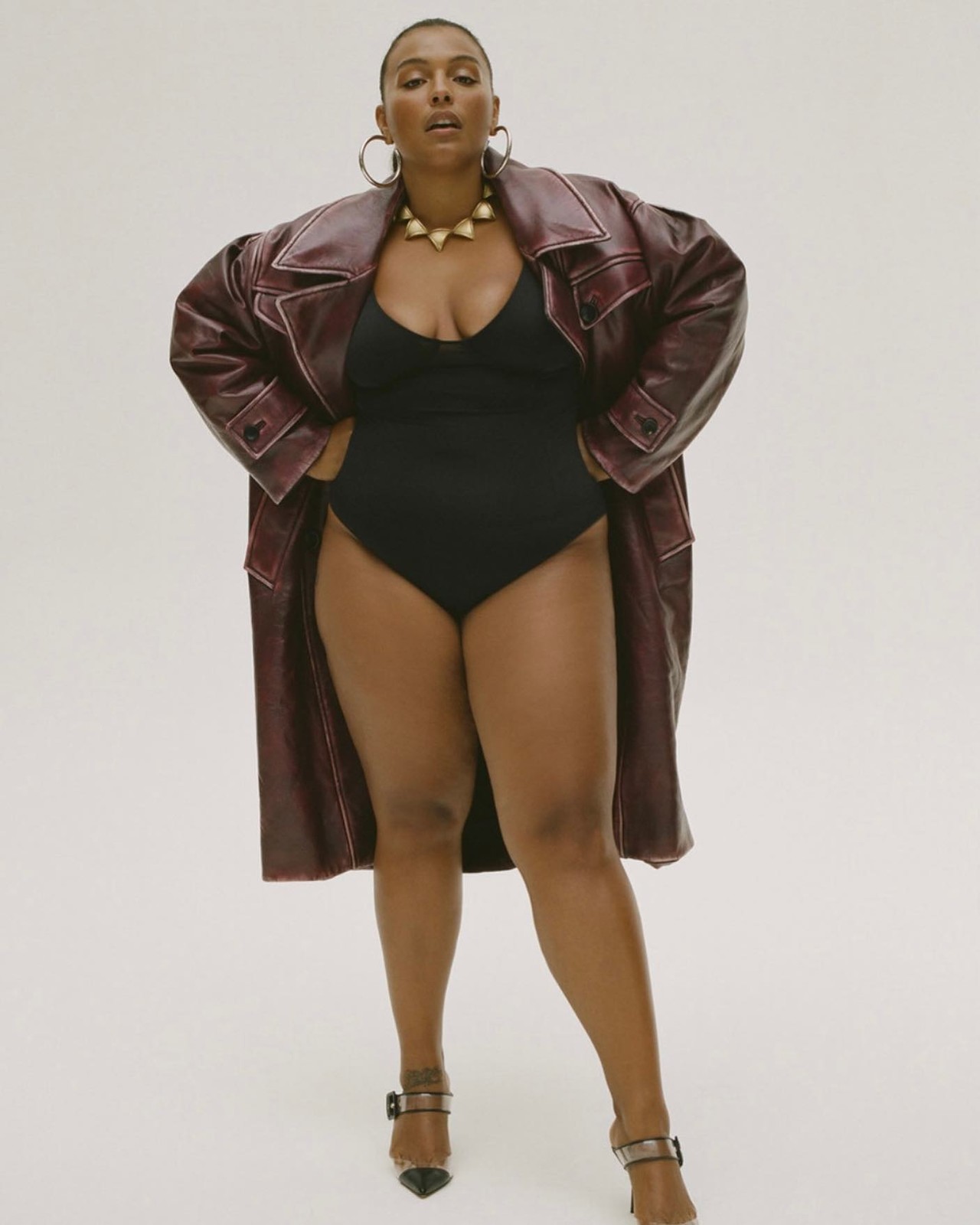 Woman poses in a black bodysuit, maroon trench coat, and heels against a white backdrop, showcasing fashion and studio photography.