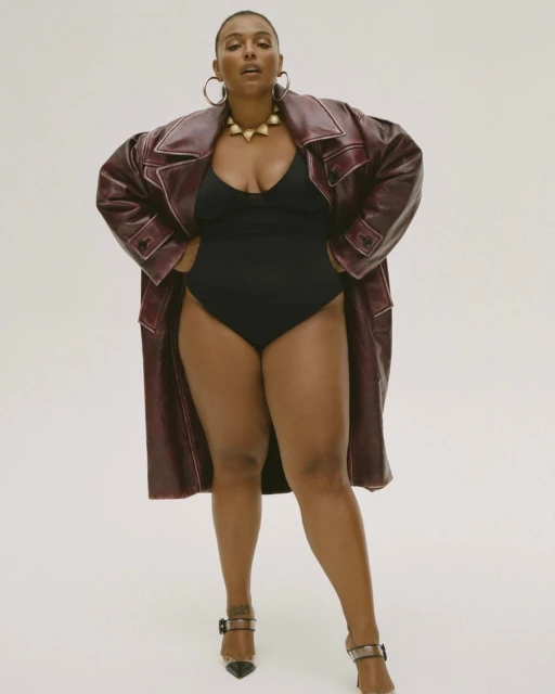 Woman poses in a black bodysuit, maroon trench coat, and heels against a white backdrop, showcasing fashion and studio photography.
