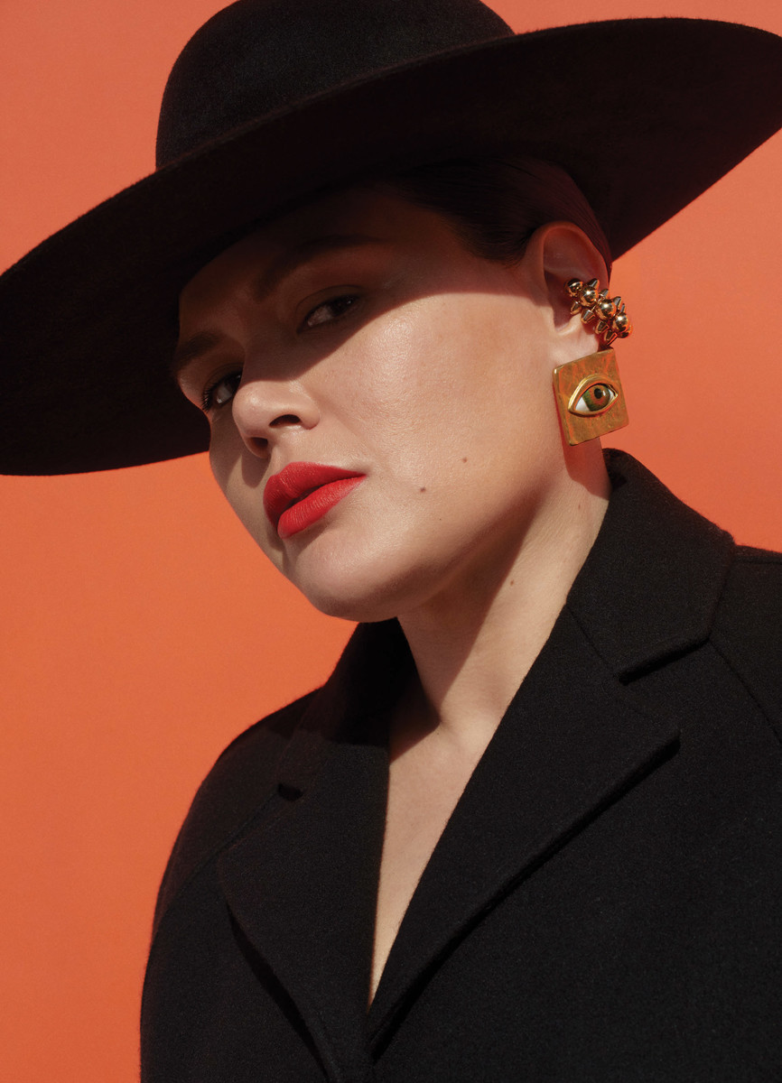 Portrait of a woman with red lipstick wearing a black coat and hat with orange background and eye-shaped earrings.