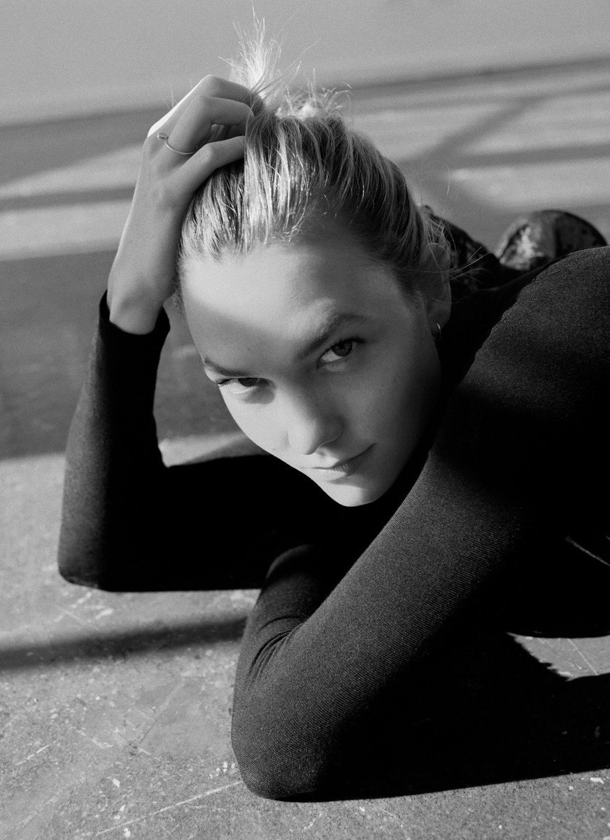 Monochrome portrait of a young woman with her hands behind her head, lying on a grey floor, looking directly into the camera.