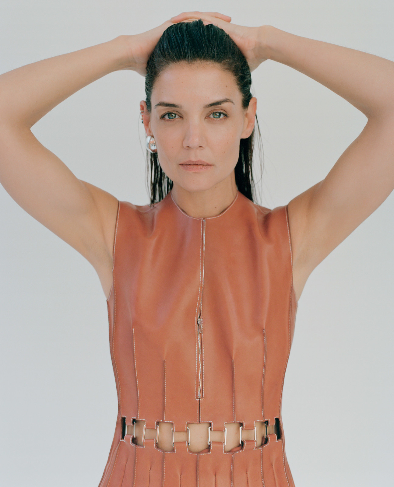 Close-up portrait of a brunette woman with wet hair and a sleeveless dress, hands above head, in a brightly lit photo studio setting.