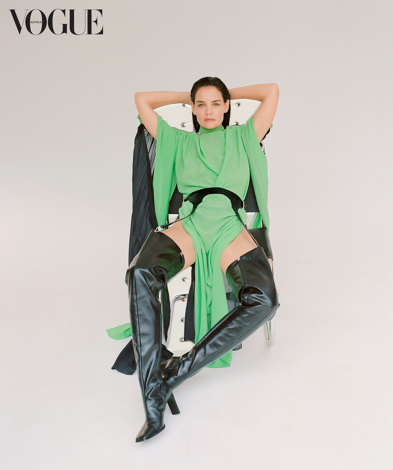 Katie Holmes seated in a chair wearing a green dress and black leather thigh-high boots against a plain background