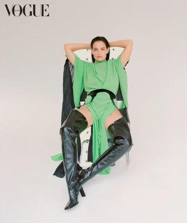 Katie Holmes seated in a chair wearing a green dress and black leather thigh-high boots against a plain background
