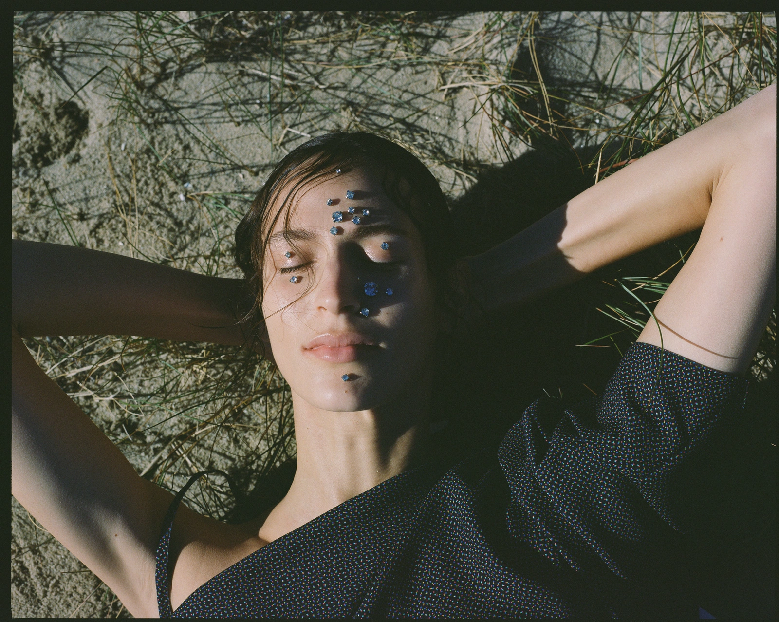 Close-up portrait of a woman lying on sandy ground with jewels adhered to her face, photographed by Bec Parsons.
