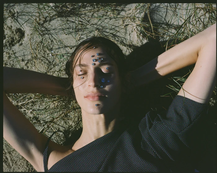 Close-up portrait of a woman lying on sandy ground with jewels adhered to her face, photographed by Bec Parsons.