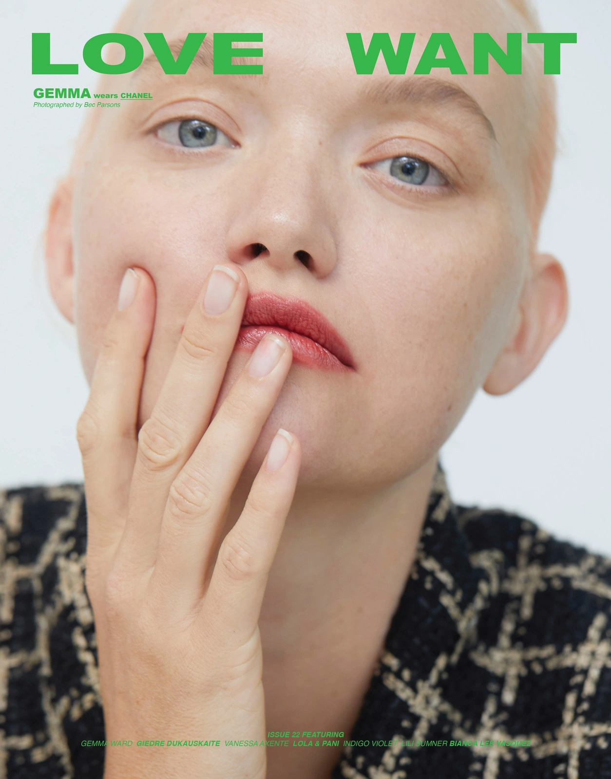 Close-up portrait of a model with short blonde hair and blue eyes touching her lips against a plain white background