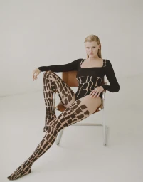 Model with slicked-back blonde hair seated on a tubular steel chair wearing a black bodysuit and patterned tights against a white background
