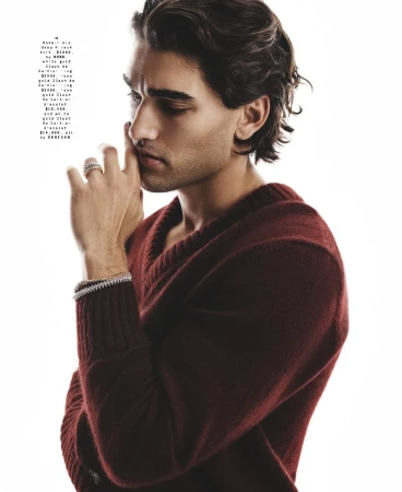 Man wearing a burgundy v-neck sweater with layered metal rings and bracelets posing against a white background