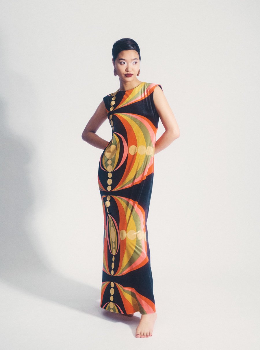 Elegant woman posing in a retro-inspired, patterned maxi dress against a stark white background in a studio setting, full-length shot.